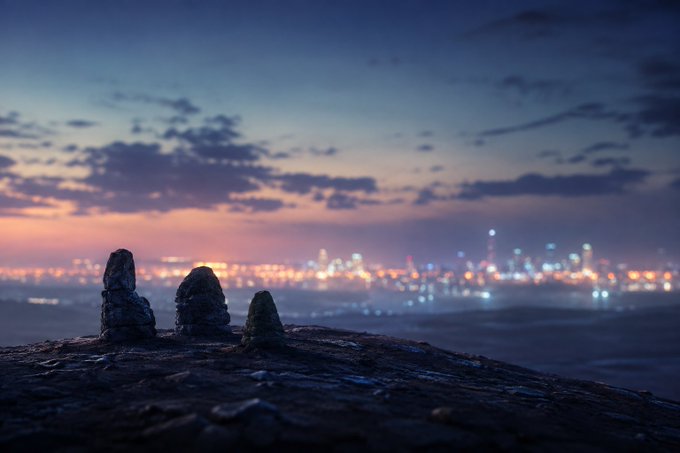Three small rock cairns stand on a dark, weathered surface in the foreground, overlooking a blurred city skyline glowing with scattered lights at twilight. The image's theme of quiet endurance amid a distant, illuminated cityscape mirrors the post's message about overlooked XRPL meme communities like $FUZZY, $ARMY, and $PHNIX persisting with modest liquidity while remaining largely unnoticed by the broader crypto world.