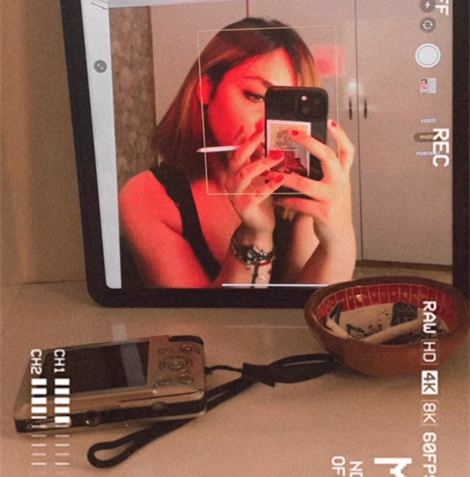 A person takes a mirror selfie with a phone, with a vintage flip phone and a bowl of keys resting on the counter in the foreground. The image is displayed through a recording app interface showing "REC," "VIDEO," "PHOTO," "PORTRAIT," and recording quality options like "RAW," "HD," "4K," "8K," "68FP," which ties into the post's theme of capturing life's everyday moments ("The Bitter Side of Life") as part of a digital NFT auction on Manifold priced at 0.12 E.