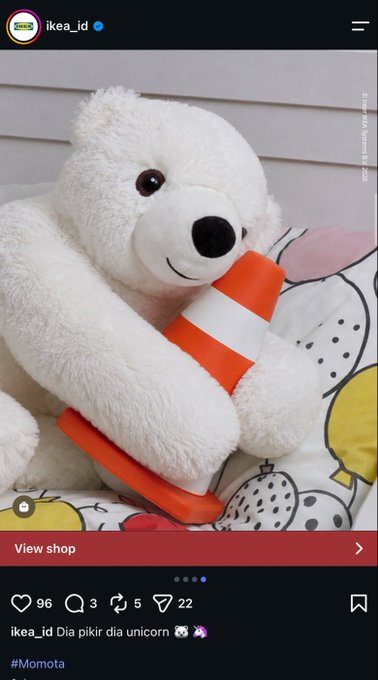 An Instagram post screenshot from 'ikea_id' showing a white teddy bear holding an orange and white striped cone, with the caption 'ikea_id Dia pikir dia unicorn' and hashtags including #Momota.