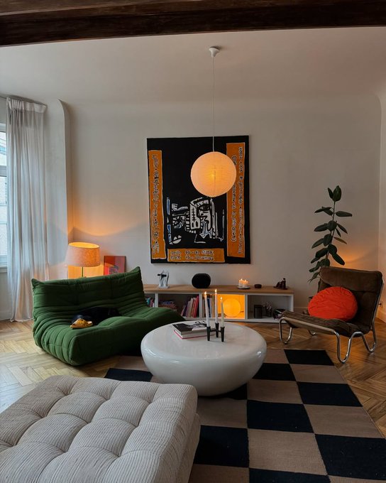 A cozy living room with a green Togo sofa, a white round coffee table, a checkered rug, a black and yellow Arabic calligraphy tapestry, and modern lighting including a paper lantern pendant.