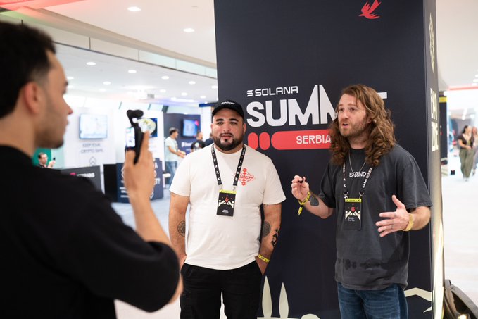 Two men wearing lanyards pose in front of a "SOLANA SUMMIT SERBIA" backdrop while a third person films them with a phone, capturing an on-the-floor moment from the crypto conference highlighted in the post.
