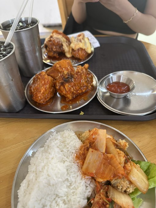 A meal tray featuring Korean fried chicken with sauce, a small dish of red dipping sauce, rice, and kimchi, alongside metal tumblers.