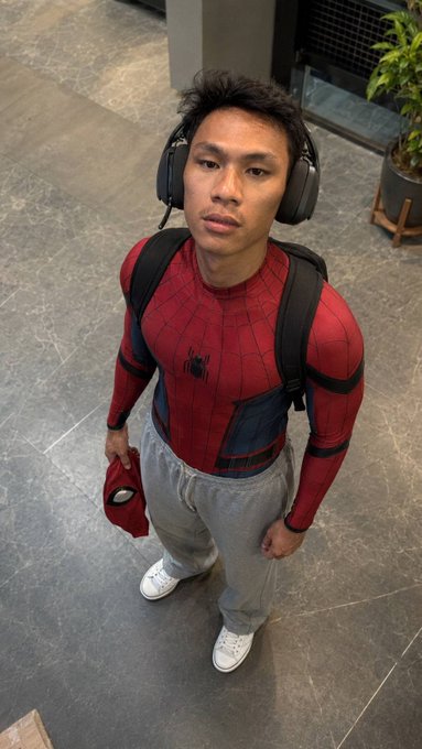 A man dressed in a Spider-Man costume and large headphones stands holding a mask, wearing grey sweatpants and a backpack.