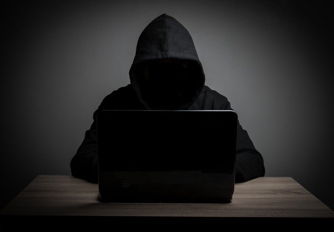 A hooded, faceless figure sits at a laptop in a dark setting, visually representing an anonymous hacker. The imagery reinforces the post about a $3 million frontend exploit on Polymarket, symbolizing the unidentified attacker behind the supply-chain attack.