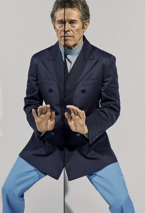 An older man in a dark double-breasted blazer and light blue trousers poses in a crouched, symmetrical stance with both hands raised in front of a white background.