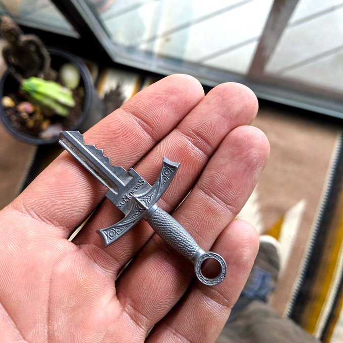 A small metal sword-shaped pendant or charm held in a person's palm, featuring a textured handle with a ring at the end and decorative engravings on the crossguard.