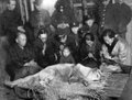 A vintage black-and-white photograph shows a group of people gathered solemnly around a deceased dog lying on a bed, evoking the legendary story of Hachiko, the loyal Akita who faithfully waited for his owner at Shibuya Station.