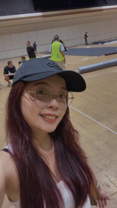 A woman wearing a black "ALO" cap and glasses takes a selfie in an indoor arena where crew members in high-visibility vests are setting up flooring and rolling out mats, capturing the behind-the-scenes preparations on setup day for the Malaysia Badminton World Championships.