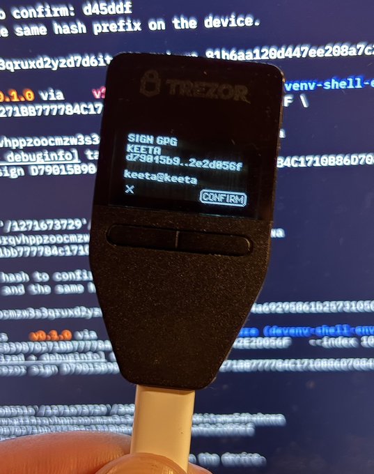 A hand holds a Trezor hardware wallet in front of a computer screen displaying code and terminal output. The Trezor's small display shows "SIGN GPG" at the top, followed by "KEETA," a partial hash "d79d45b9..2e2d856f," the recipient "keeta@keeta," and an "X" with a highlighted "CONFIRM" button, indicating it is awaiting user approval to sign a GPG signature for the Keeta blockchain.