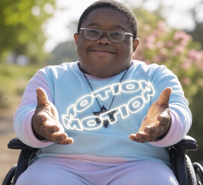 A young man with glasses wearing a light blue 'MOTION' sweatshirt sits in a wheelchair outdoors with his hands extended toward the camera.