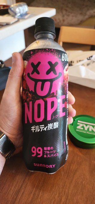 A hand holding a black bottle of Suntory 'NOOPE' sparkling carbonated drink with a pink smiley face design, with a green ZYN container on the table beside it.