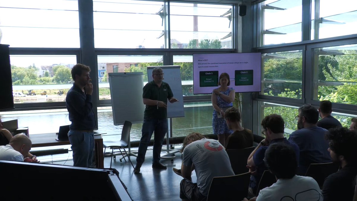 The image shows a group of people gathered around two presentations happening simultaneously. One presentation has a man standing behind a whiteboard with a green board, while the other presentation features a woman standing next to a large screen displaying information about EEZs. The audience appears engaged and interested in both presentations.