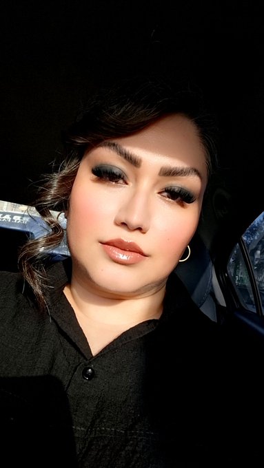 A selfie of a woman wearing heavy makeup with dark, dramatic eyelashes and glossy lips, taken inside a vehicle.