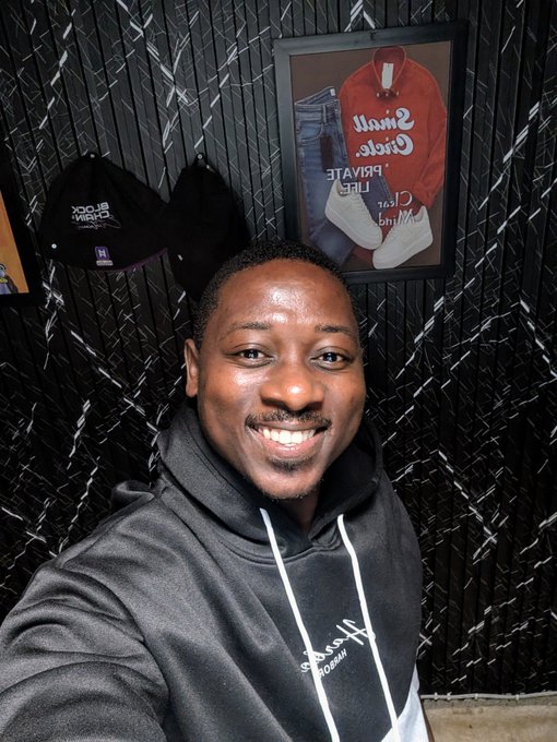 A smiling young man takes a selfie in front of a framed "Santa Cruz Private Club" vintage display featuring a red "Santa Cruzed" hoodie and jeans, with a black "BLOCK" cap mounted on the dark textured wall beside it. The image complements the upbeat "GM Folks" Friday post, radiating a cheerful, weekend-ready vibe.