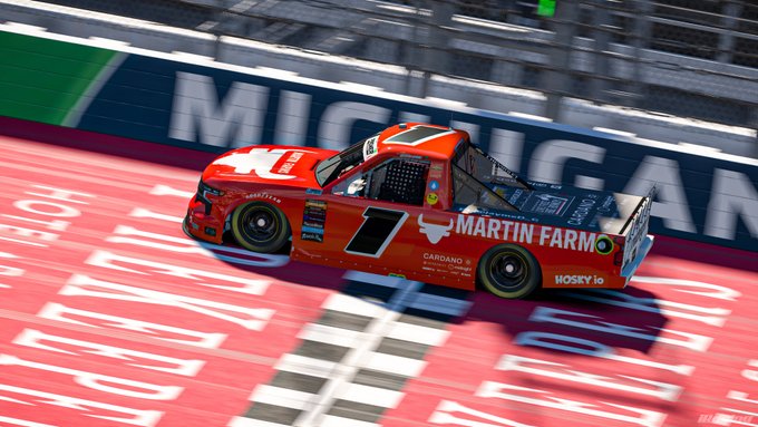 The image features a red truck racing along a track, with the word "Martin Farm" displayed on its side. The truck appears to be driving at high speed, possibly during an important race or event. In the background, there are several people standing around, likely spectators or support staff for the race. One person is holding a sign that says "@MISpeedway", which could indicate the location of the racing event.