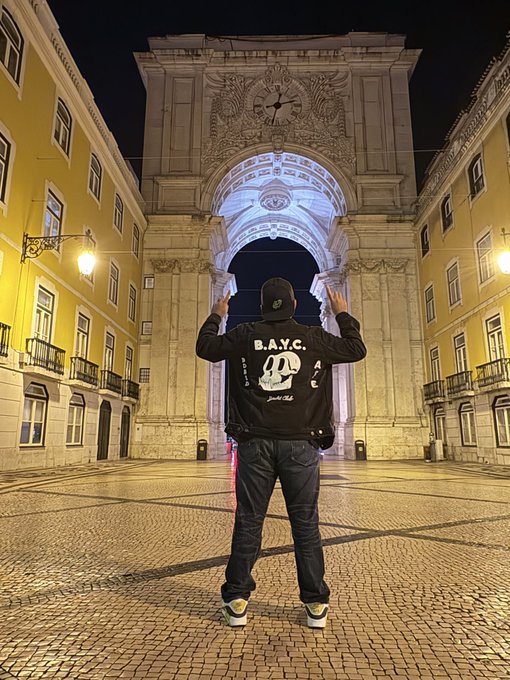 Nighttime photo of someone wearing a "B.A.Y.C." (Bored Ape Yacht Club) jacket standing in front of the illuminated Arco da Rua Augusta in Lisbon, Portugal.