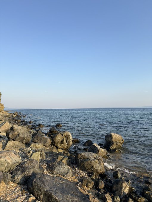 A peaceful coastal scene shows a rocky shoreline leading into a calm, blue ocean under a clear sky.