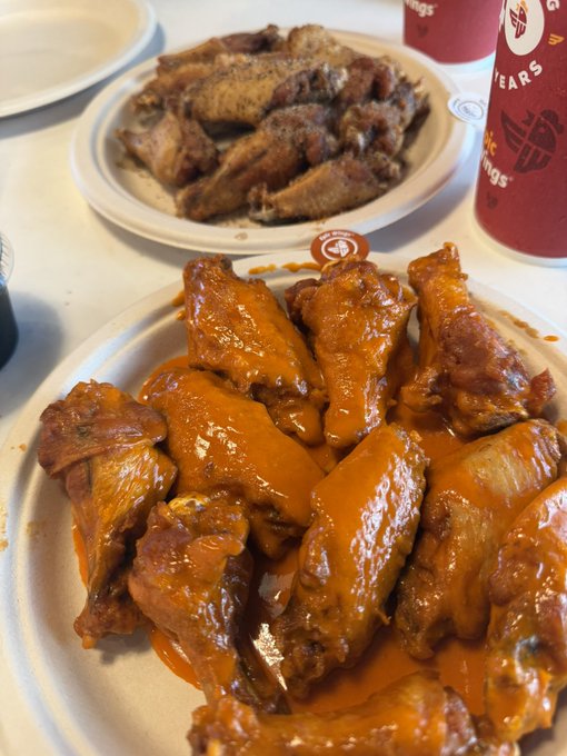 A close-up shot of two plates of chicken wings on a table, with the foreground plate featuring wings coated in a bright orange buffalo-style sauce and the background plate holding darker, seasoned wings, alongside red cups bearing a wings logo.