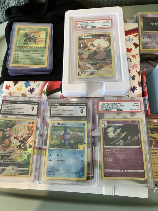 Several graded Pokémon trading cards in protective slabs are displayed on a colorful patterned surface, including PSA 9 cards like Eevee, Mudkip, and Aerodactyl, along with a CGC 8 and 9 graded card.