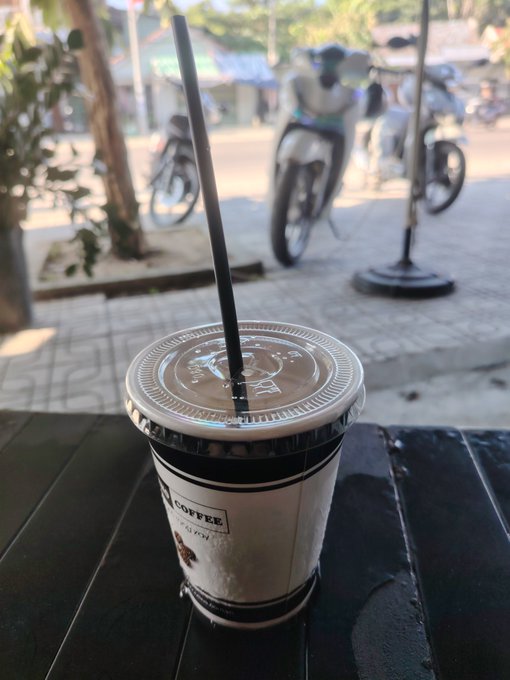 An iced coffee with a black straw sits on a dark wooden table at a street-side café, with motorcycles parked on the road in the background.