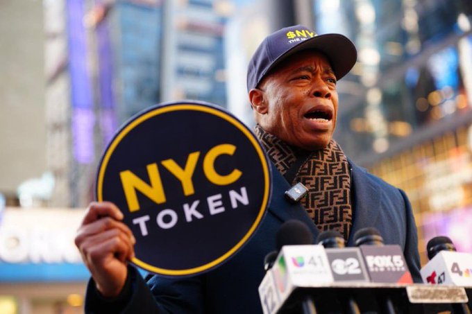 A man wearing a '$NYC TOKEN' cap holds a circular 'NYC TOKEN' sign at a press conference with multiple microphones.