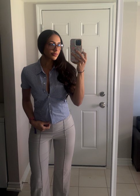 A mirror selfie of a young woman with long dark hair and glasses, wearing a light blue short-sleeve button-up shirt and white pants, standing in front of a white door.