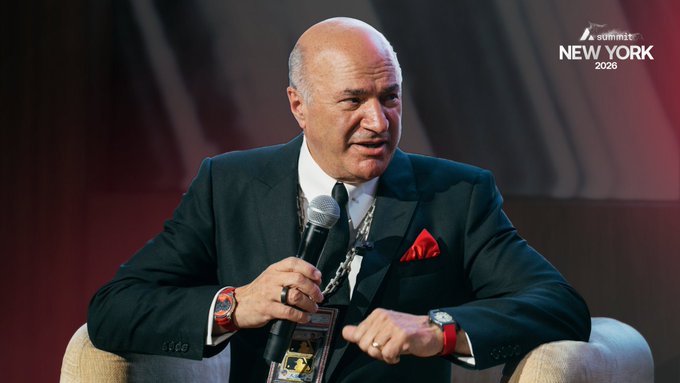 A bald man in a dark green suit and red pocket square speaks into a microphone at an event, with a lanyard around his neck and a 'SUMMIT NEW YORK 2026' logo visible in the upper right corner.