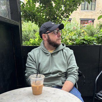 The image features a man sitting at an outdoor table, wearing a black cap and glasses, with his hand resting on his chin as he looks off into the distance. On the table next to him, there's a drink in a plastic cup. The background includes some greenery from trees and bushes, along with a building visible behind them.

The post is titled "Web2 vs Web3 Nemo We doxxed out here I am my @doodle", which seems to be a humorous take on the ongoing debate between web browsers (Web 2) and web browsers (Web 3). The image includes elements from both sides of this debate, with the man in glasses representing one side and the post itself representing the other.