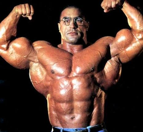 A photograph of a muscular bodybuilder posing in a double biceps front pose, showing extreme muscular development and definition against a black background.