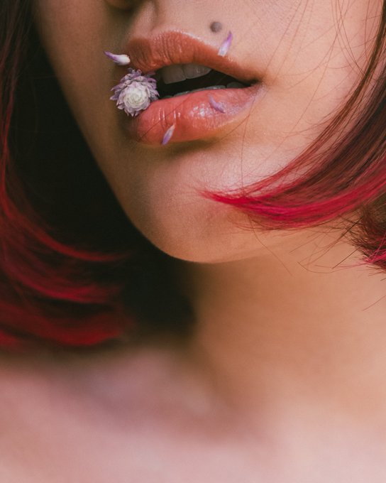 A close-up photograph of a woman's lips featuring a small purple flower decoration and purple nail-shaped accents, with red-tinted hair visible on the side.