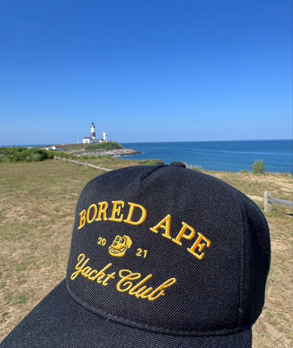 A black snapback hat embroidered with "BORED APE 20 21 Yacht Club" in gold lettering sits in the foreground against the iconic Montauk Lighthouse and ocean backdrop, perfectly tying together the BAYC NFT theme with the scenic Long Island location.