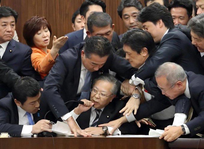 A chaotic scene in what appears to be a Japanese parliament showing several men in suits involved in a physical altercation, with one man in the center appearing to be restrained by others.