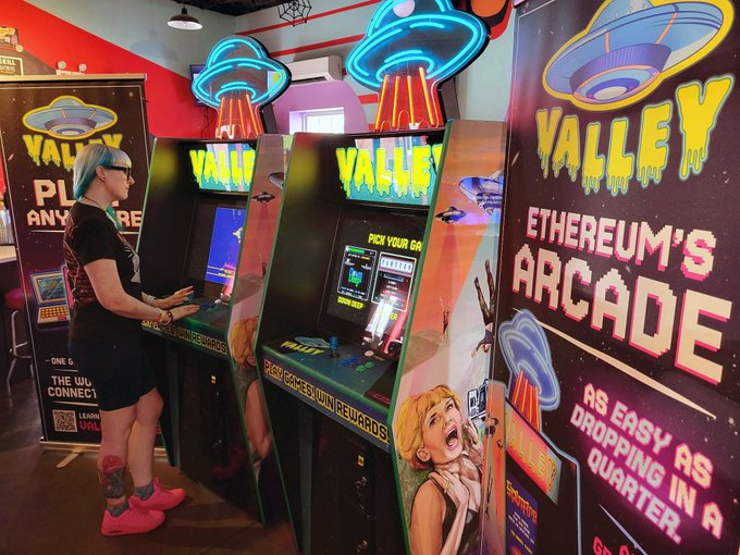 The image shows a woman standing next to two video game arcades, one of which has a sign that says "Valley Arcade" above it. The other arcade appears to be an old-fashioned arcade with neon signs and a poster for an event called "ETHConf!". The woman is wearing a black shirt and pink shoes while looking at the games in front of her.

The image also contains text elements, such as the words "game on" and "Valley Arcade Zone", which suggest that there might be a special event or promotion happening at this arcade.