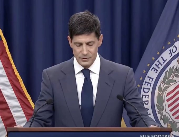 A man in a blue suit and tie stands at a podium looking down, with a US flag on the left and a Federal seal on the right; the image is labeled 'FILE FOOTAGE' in the bottom right corner.