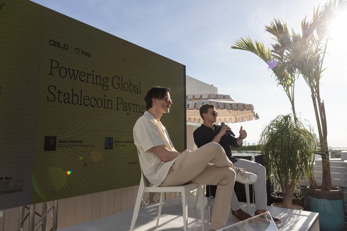 The image shows two people sitting on chairs outside a building with a large screen behind them displaying information about Celo, an Ethereum-based stablecoin project. The text on the screen reads "At @consensus2026, @Bridge announced support for Celo-native USDC. Ready to build global stablecoin payment infrastructure? Fiat → USDC on Celo, now via one API". One person is sitting in a white chair and the other is sitting in a black chair. They are both looking at the screen attentively, possibly discussing or planning something related to the project.