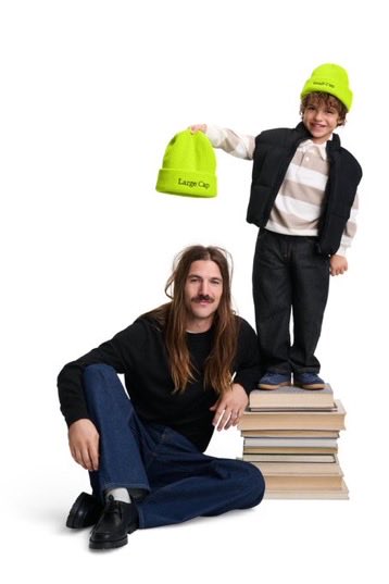 A young boy in a yellow beanie and vest holds up a bright green beanie labeled "Large Cap" while standing on a stack of books, next to a seated long-haired man in a black sweater and jeans.