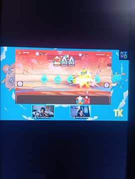 A blurry photo of a television screen displaying a colorful video game with a fighting interface, showing player avatars at the top and character selection boxes at the bottom.