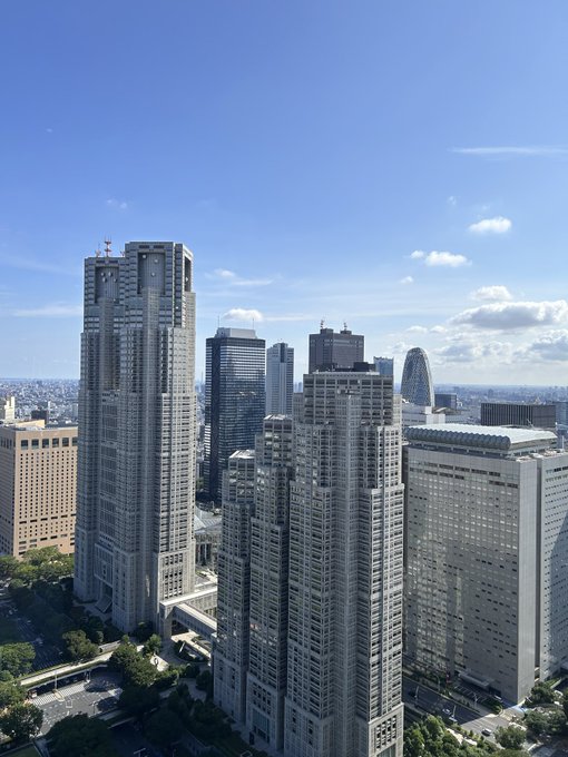 A daytime cityscape showing several tall skyscrapers, including one that resembles the Tokyo Metropolitan Government Building, under a blue sky with scattered clouds.