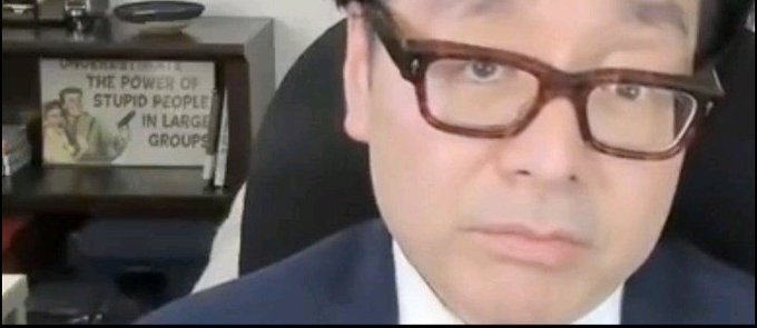 The image features a man wearing glasses with brown frames sitting behind a desk. The background of the image shows an office setting, which includes a bookcase filled with books. A sign can be seen hanging above the man's head, but its content is not visible in this particular frame. The main focus of the image is on the man and his surroundings, creating a sense of depth and context for the viewer.