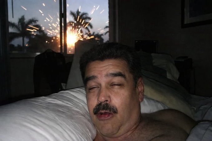 A shirtless man with a mustache lies in bed with his eyes closed while sparks fly outside the window behind him.