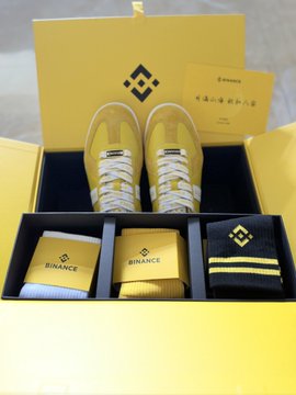 A yellow promotional gift box containing yellow sneakers, socks, and other items, all branded with the "BINANCE" logo.