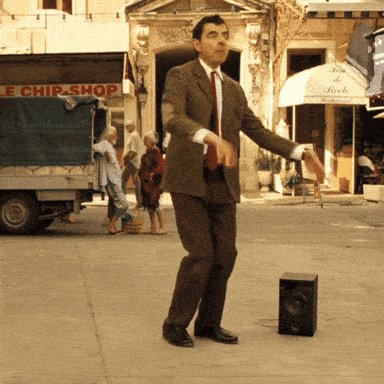 The image features an actor dressed in a suit and tie standing on a street with his arms outstretched as if dancing, holding a microphone or speaker. The background includes a building and some people walking by. There are also two trucks visible in the scene - one near the left side of the image and another further back to the right. A sign for "chip shop" is present, indicating that this could be an outdoor market area.