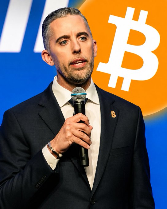 A man in a dark suit with a Bitcoin lapel pin speaks into a microphone, standing in front of a backdrop featuring a large Bitcoin logo, likely at a crypto-related conference.