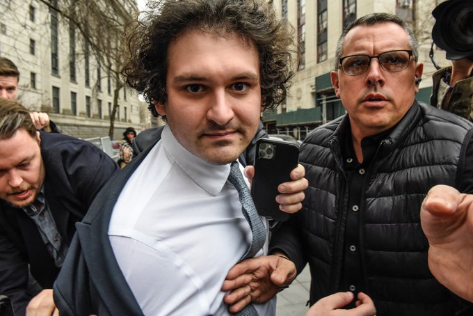 A photograph showing Sam Bankman-Fried in a suit and tie being escorted by men in a crowded urban setting.
