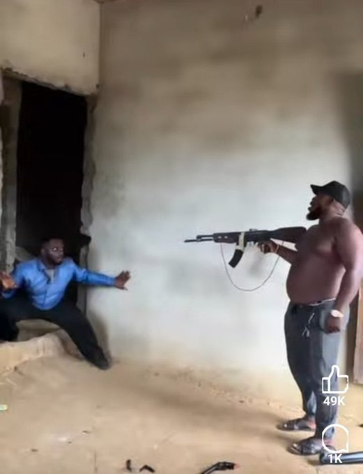 A screenshot from a video shows a shirtless man in a cap pointing a rifle at another man in a blue shirt who is cowering against a wall in an unfinished room, with 49K likes and 1K comments visible.