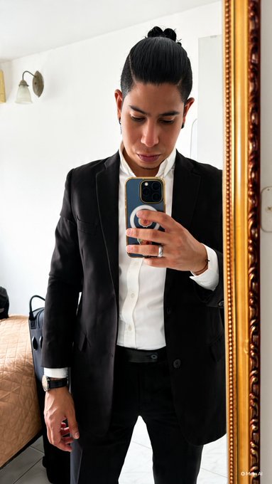 A man with a man bun in a black suit takes a mirror selfie holding a phone.