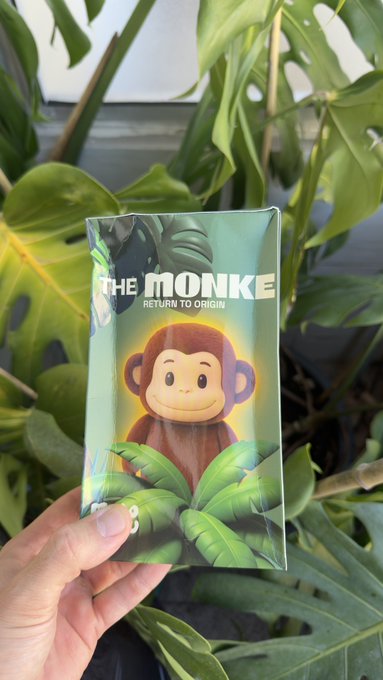 A hand holds a blind box for 'THE MONKE - Return to Origin' featuring a cartoon monkey face, photographed against green Monstera plant leaves.