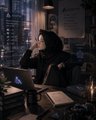 The image shows a person sitting at a desk with a laptop computer, wearing a black hoodie or headscarf. The background features a window overlooking a cityscape at night, which adds an urban atmosphere to the scene. There are also several books on the desk and a candle in the room, creating a cozy and intellectual ambiance.