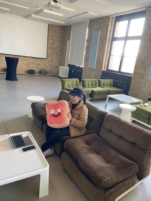 A person wearing a bucket hat and glasses sits on a brown couch in a modern lounge area with exposed brick walls, holding a pink cartoon-faced plush pillow, with a laptop and phone on a white coffee table.