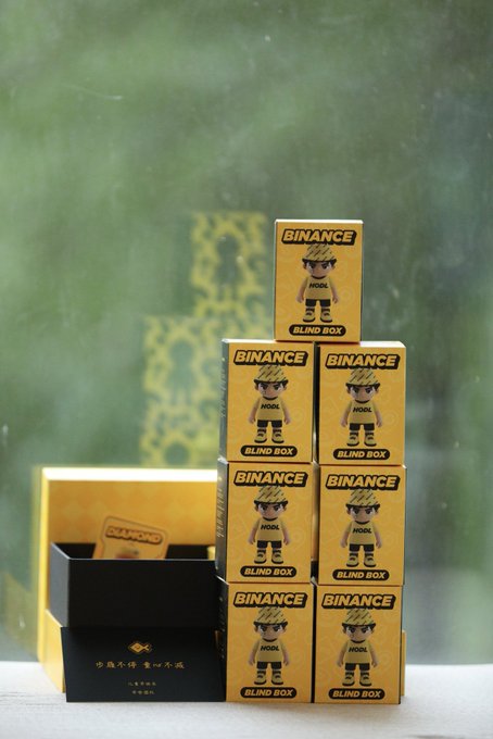 The image features a stack of seven bins, with one bin labeled "binance" and another labeled "blind box", both of which are yellow. The bins appear to be made out of cardboard or paperboard material. In addition to these bins, there is also a small black box placed on the table next to them.